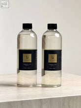 Two clear glass bottles with black caps and gold labels containing amber fie refill for lifestyle home square reed diffuser