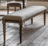 Artisan dining bench seat with light gray upholstered cushion and turned wooden legs in a rustic finish