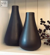 Two black glass vases with a matt finish and tapered teardrop shape sit side by side on a shelf