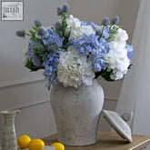 A rustic white ceramic stone ginger jar vase with textured finish holding blue and white artificial hydrangeas