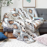 A cozy boho woven throw with earthy tones white base and geometric brown black and teal patterns with fringed edges