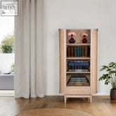 Beige chester cane bookcase with woven cane sides mango wood drawer and expansive shelves holding books and pink vases