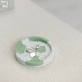 A chunky trinket dish with a round speckled concrete design and mint green geometric accents holding silver jewelry
