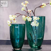 Two dark green mouth blown glass vases from dutz collection with ribbed texture holding white blossoms on branches