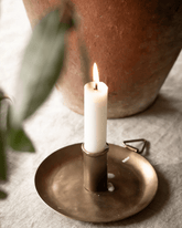 A white dinner candle burning in a hand forged brass candle holder with a circular base and metal clip nearby