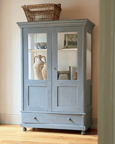 Hand painted victorian cupboard in rustic gray with glass doors interior shelves and a wicker basket on top