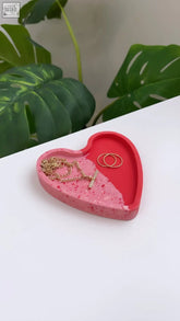 Pink and red heart trinket dish with a speckled resin texture holding gold rings and a delicate chain necklace