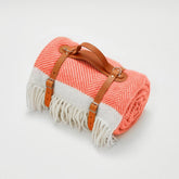 A coral and white herringbone wool picnic blanket with tan leather straps and fringed edges perfect for outdoor use
