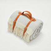 A soft cream and light gray herringbone wool picnic blanket with long fringe and a tan leather strap handle