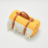 A rolled yellow and white herringbone wool picnic blanket with tan leather straps perfect for outdoor use