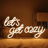 A warm white cozy neon light sign that reads lets get cozy in cursive for bedroom bar or cafe walls