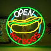 A glowing neon sign shaped like a hot dog with open hot and hot dogs words in white red green and yellow tubing for wall decor