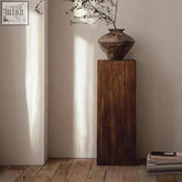 A rustic earth-toned ceramic vase with textured surface on a dark solid wood pedestal showing natural grain on kumo side table