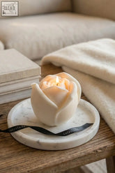 A creamy white large valentine’s day rose shaped candle with a lit wick on a marble tray with interlude ribbon
