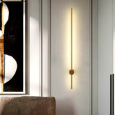 Gold wall light with sleek vertical bar and circular base emitting warm light perfect for modern bedroom lounge