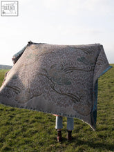 A soft gray woven nouveau blossom blanket with intricate floral patterns and fringed edges draped over a person in a grassy field