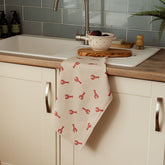 A cream linen tea towel with red lobster prints hanging over a wooden countertop edge perfect for kitchen use