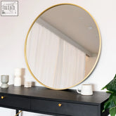 A 90cm round wall mirror with a sleek gold metal frame sits on a dark wooden console table for stylish decor