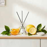Clear glass revitalising diffuser 100ml with black reeds on wood surrounded by bright orange and lemony litsea for consistent scent