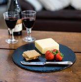 Round slate cheese board with pale yellow cheese, red strawberries, golden crackers, and silver butter knife