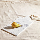 A small white marble chopping and serving board with gray veining and rope handle holding a golden pear
