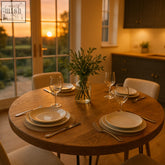 A clear glass vase with greenery sits on a solid oak round dining table with hairpin legs and a wooden finish