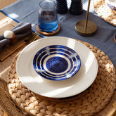 A hand painted stripe ceramic side plate with blue and white concentric circles on a woven placemat