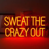 A glowing yellow led neon sign with bold letters neon spelling sweat the crazy out for gym and man cave decor