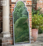 A tall arched vintage grey metal outdoor mirror with a sleek silver frame against a brick wall