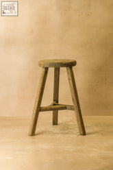 Vintage rustic elm wood stool with round weathered seat and four splayed legs joined by a central crossbar