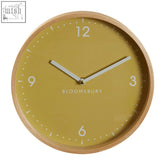 Vitus small yellow wall clock with a round light wood frame, mustard yellow face, and white hands and numerals on yellow wall