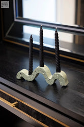 A sleek wavy candlestick concrete candle holder with a speckled green finish holding three tall black twisted candles for luxe home decor