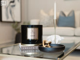 Black cylindrical xl candle with amber fie scent and gold rimmed label sits on a glass table for cozy use