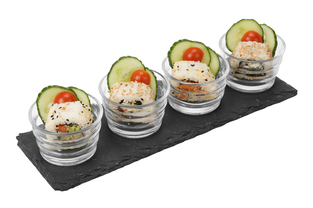 Four clear ribbed glass bowls with mini sushi rolls on a black slate tray garnished with cucumber and cherry tomatoes