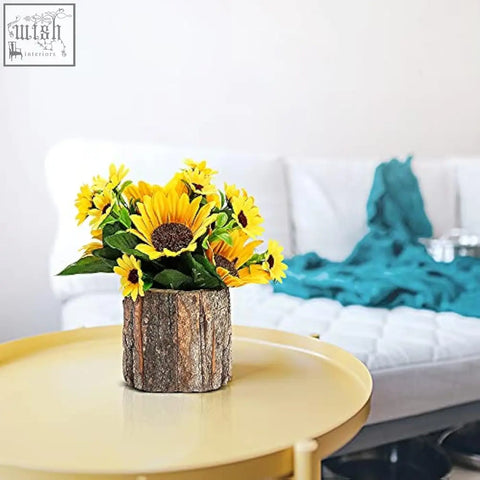 A rustic wooden log vase holding bright yellow artificial sunflowers with green leaves for indoor decor
