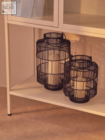 Black wire lanterns with cylindrical candles inside sitting on a white metal cabinet with glass doors