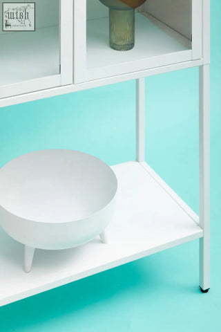 White ceramic bowl with three short legs displayed inside a white metal cabinet with glass doors