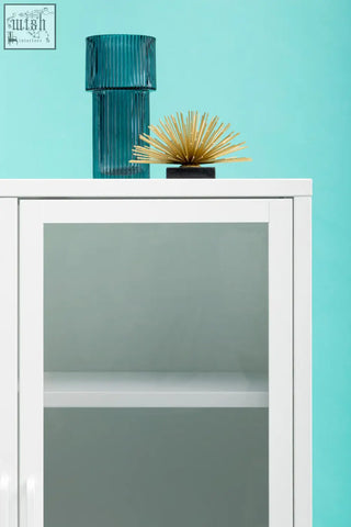 A tall teal glass vase with vertical ridges on top of a white metal cabinet with glass doors