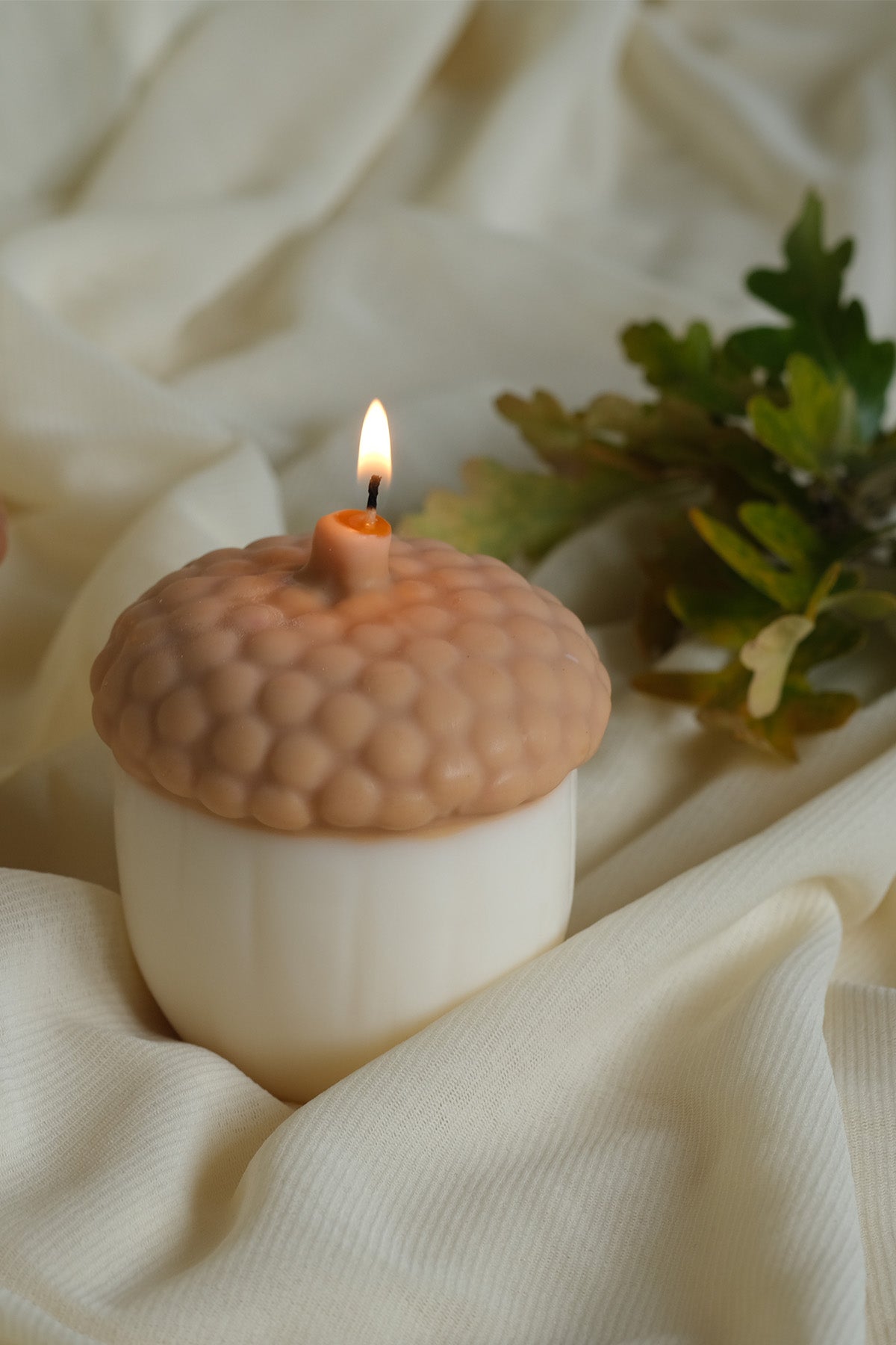 A lit cream-colored acorn shaped soy wax candle with a textured brown cap and small flame
