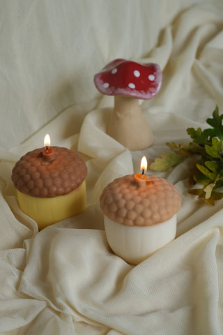 Two lit acorn shaped soy wax candles with textured brown caps and smooth yellow and white bases