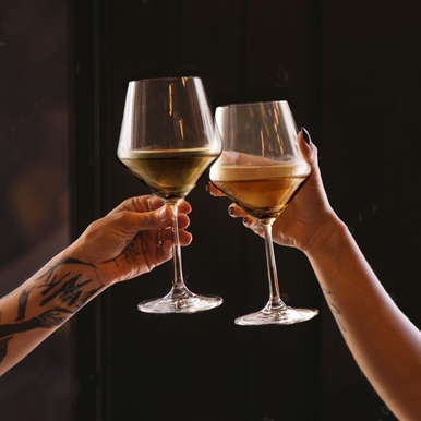 Two hands hold up clear crystal burgundy glasses filled with golden white wine against a dark background