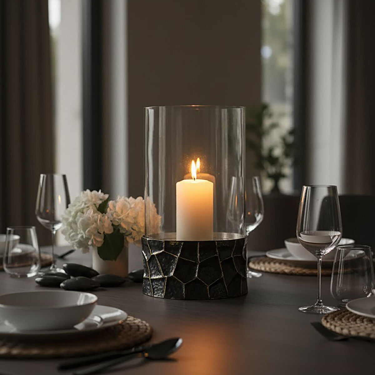 A tall clear glass candle holder with a textured black base holding a lit white pillar candle akola gold large hurricane