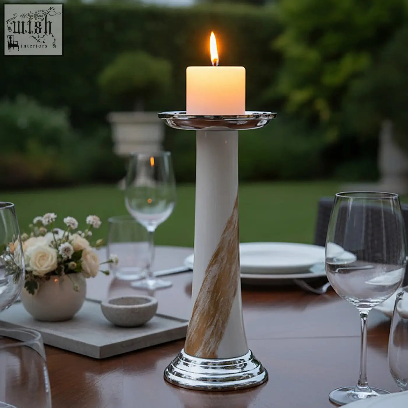 A tall elegant alma white and gold candle holder with gold swirls and an orange glowing candle on a silver rim plate