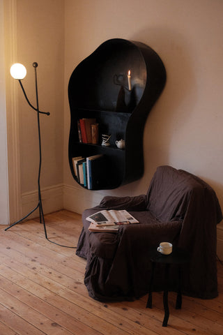 Black organic-shaped wall shelf with curved edges holding books and decor next to aluminium balancing act floor lamp