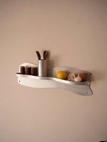 A sleek white aluminium middle shelf with a wavy metal design holding kitchen items on the wall