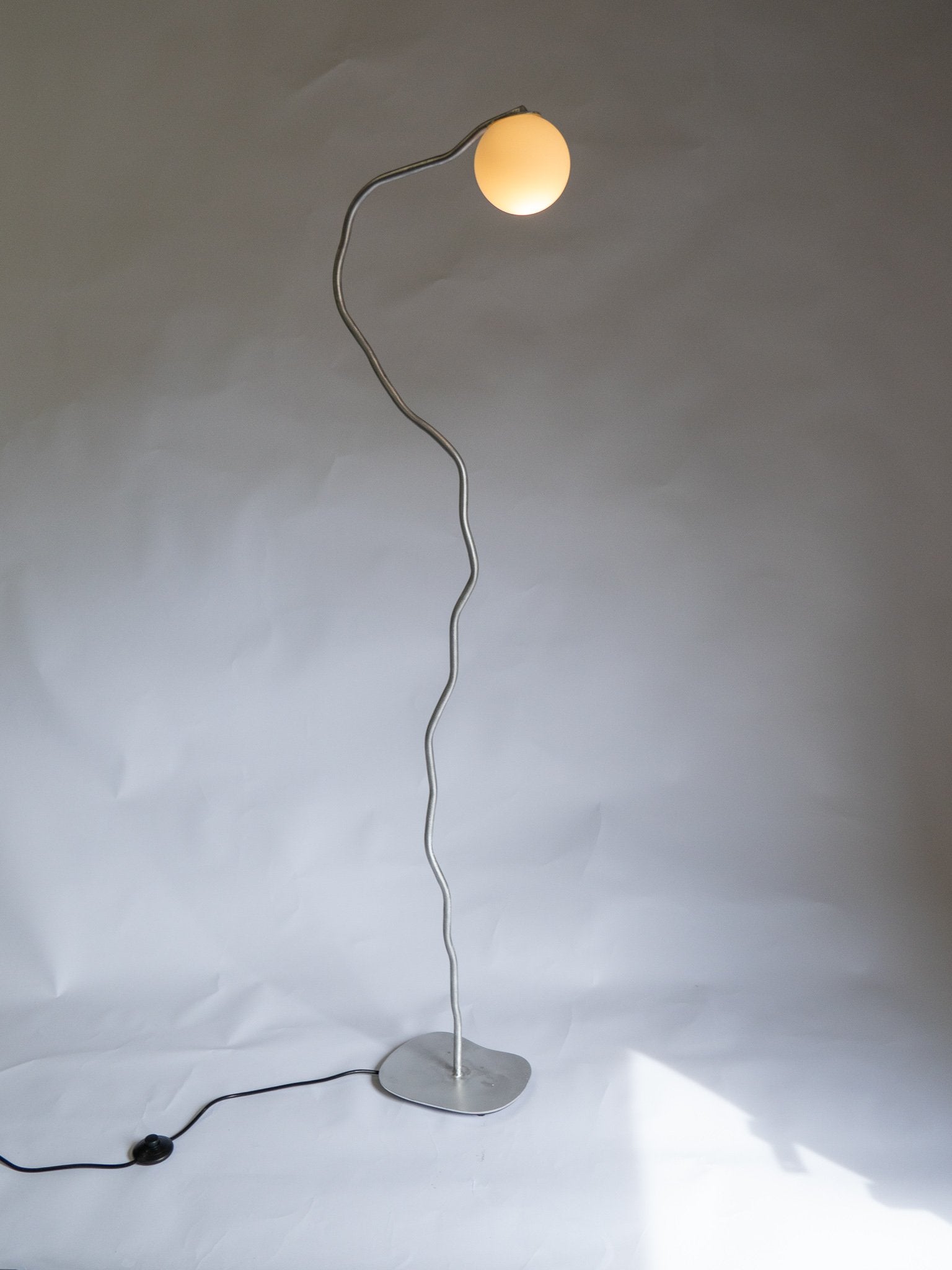 A sleek silver aluminium pea head floor lamp with a wavy metal stem and glowing spherical bulb