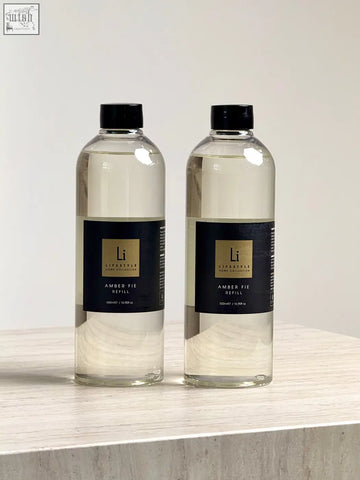 Two clear glass bottles with black caps and gold labels containing amber fie refill for lifestyle home square reed diffuser