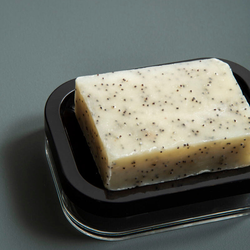 A rectangular creamy off-white soap with poppy seeds on a sleek black and transparent ando black acrylic soap dish