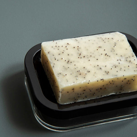 A rectangular creamy off-white soap with poppy seeds on a sleek black and transparent ando black acrylic soap dish