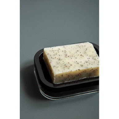 A rectangular off-white soap with dark speckles sits in an Ando black acrylic soap dish with transparent trim